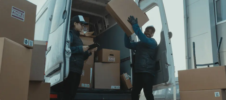kineto Two delivery workers unload cardboard boxes from a van. One person is holding a clipboard and checking boxes, while the other lifts a large box. Several packages are stacked inside and around the open van.