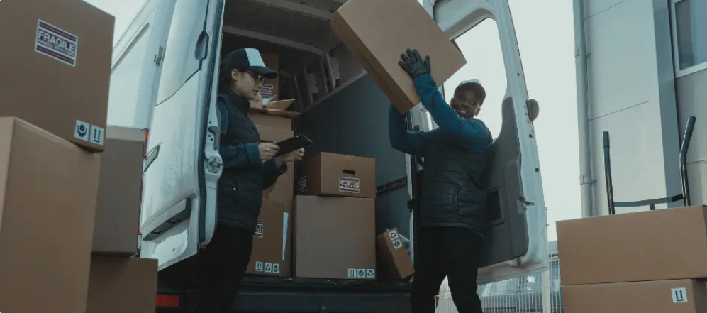 kineto Two delivery workers unload cardboard boxes from a van. One person is holding a clipboard and checking boxes, while the other lifts a large box. Several packages are stacked inside and around the open van.